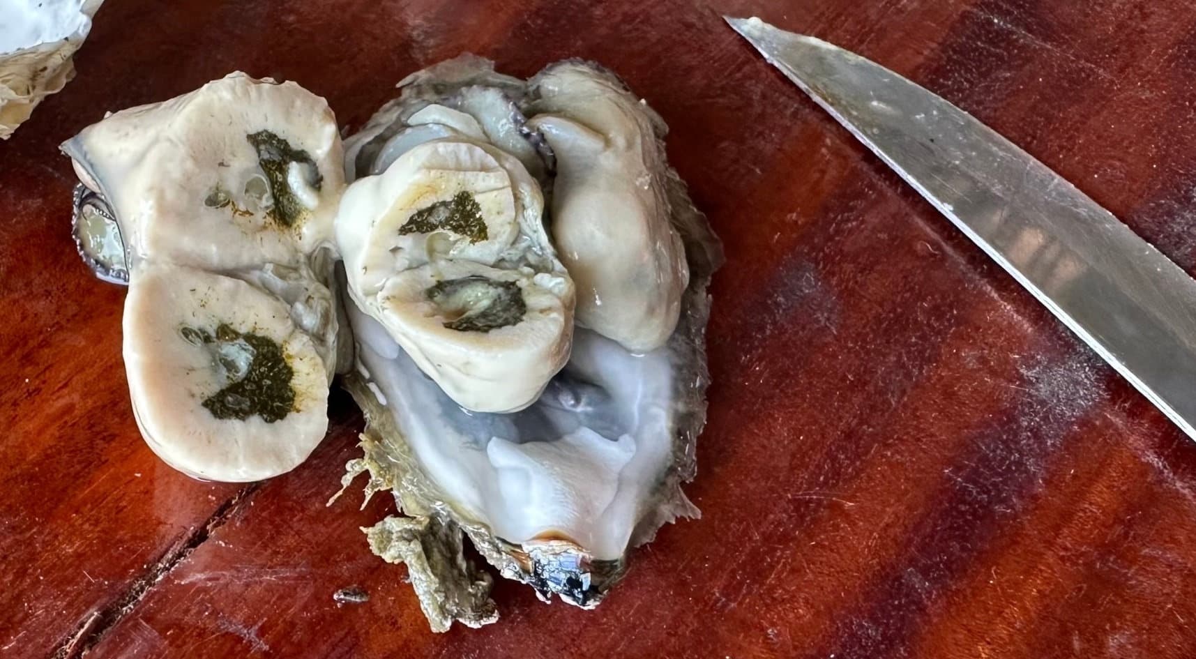 A shucked oyster cut in half on a wooden board, showing its internal cross-section, with a knife alongside.