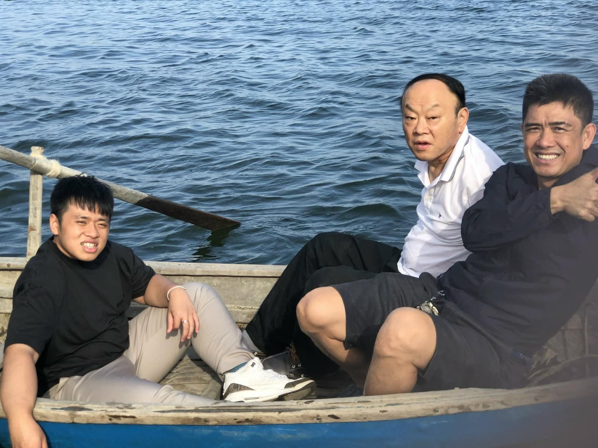 Three men sitting in a small wooden boat on open water, looking towards the camera. Takayoshi is on the left in a black shirt; Mr. Kan is in the middle in a white shirt.